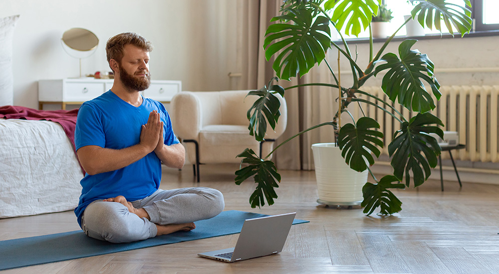 Person in bright blue tshirt sitting cross-legged on yoga mat in yoga pose with laptop open in front of him