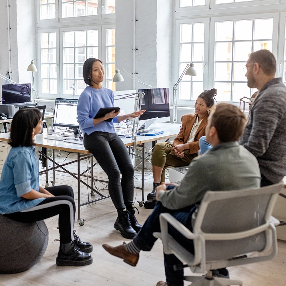 Woman perched on a corner of a computer desk explaining a concept to four people seated around her