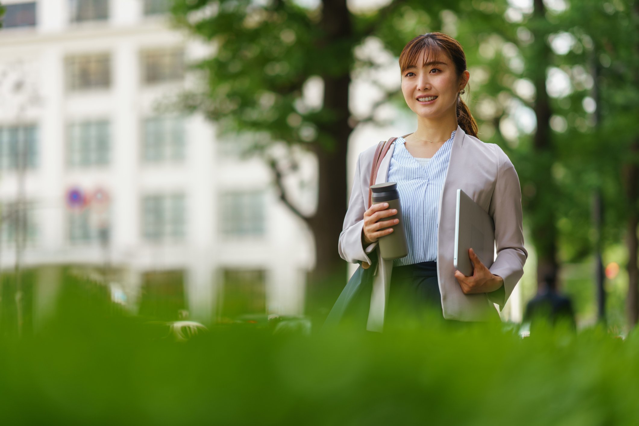 Corporate-woman-smiling-walking-with-coffee--and-laptop-through-park