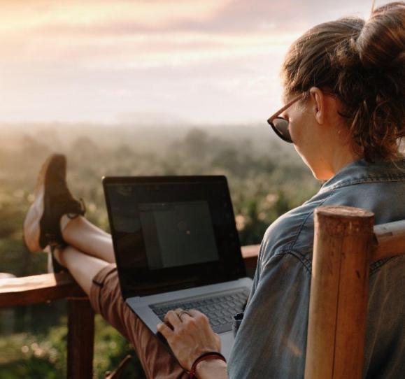 Light-skinned woman working outside with a view of a forest of trees. She sits with her feet up on railing, holding a laptop in her lap