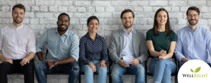 Six people sitting on a bench against a white brick wall, facing the camera and smiling