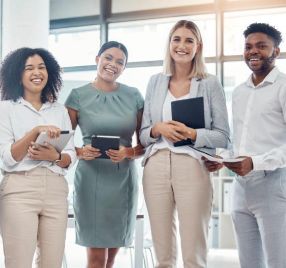 Four professionals, three female and one male, holding devices, standing in business setting, smiling at the camera