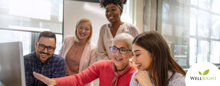 four people look at a computer monitor while an older woman with short white hair and black glasses speaks to what she is demonstrating