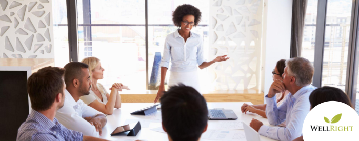 Dark-skinned woman in white stands at head of conference room table, presenting to 7 other HR professionals