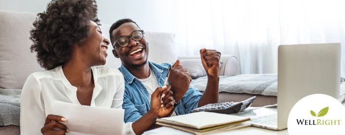 Two laughing adults, sitting in front of their sofa, looking at a laptop, with calculator